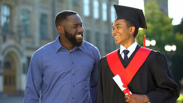 Smiling black father embracing graduating son with diploma, education degree