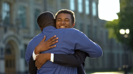 Glad parent hugging young son in suit outdoors university, prom celebration