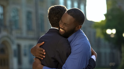 Cheerful black father embracing young son in prom suit, college graduation, joy