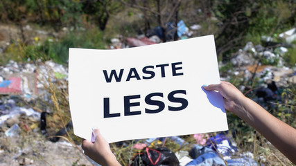 Waste less phrase on cardboard in hands against landfill background, pollution