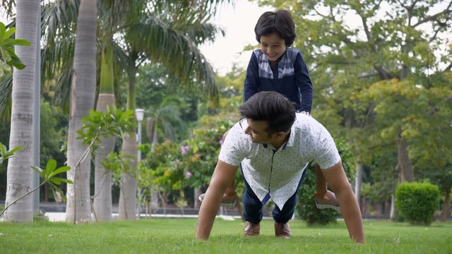 Handsome man doing push-ups with the boy on his back in casual wear - leisure time. Cute Indian kid happily enjoying the ride of pushups while sitting on his father's back - healthy lifestyle