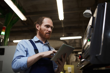 Young bearded engineer typing on digital tablet and looking at monitor of machine in the factory