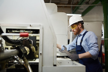 Young bearded repairman in protective workwear using wrench and working on lathe in the plant