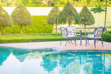 Outdoor chairs and table place nearly swimming pool with green trees and sunlight in background.