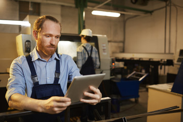 Young bearded engineer in overalls standing and working on digital tablet with woman working in the background in factory