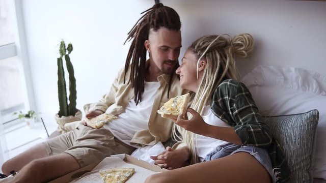 Young sweet couple has fun in bedroom, eating delivery pizza, feeding each other, lying in bed, spending weekend morning together. Caucasian guys with dreadlocks. Slow motion