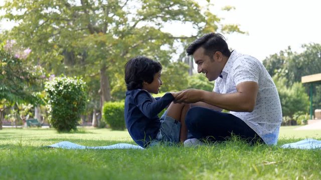 Caring Indian father helping his son while doing crunches - sports lifestyle. A young attractive man helps his kid in performing sit-ups while holding his hands and legs for healthy living - park v...