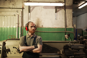 Young serious mechanic in overalls standing with arms crossed and thinking about something in warehouse