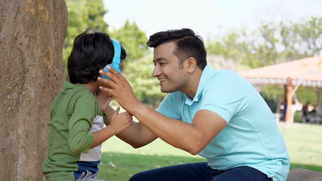 Young handsome father enjoying the weekend with his son - Indian family concept. Cute little kid happily listening to music using his Bluetooth headphones while dancing with his dad - technology co...