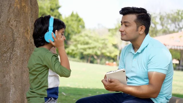Indian father and son using a tablet with blue headphones while spending time together. A cute little boy listening to music using his Bluetooth headphones while having fun with his dad - technolog...