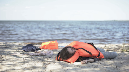 Young woman in life jacket lying on seashore, feeling unwell after shipwreck
