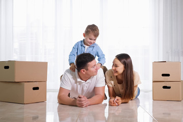 cute child sits on father back lying on light floor in new house near beautiful wife among cardboard boxes
