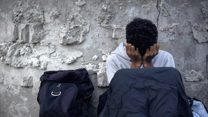 Poor afro-american refugee crying, sitting street in sleeping bag, homelessness