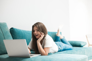 beautiful young woman having online conversation on her laptop lying on the sofa laughing