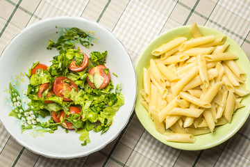 Plate with tubular pasta next to plate of tomato and cucumber salad.