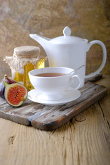 A white cup and a white teapot with tea, a jar of honey and fresh ripe figs on the cutting board on the table.