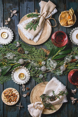 Fototapeta na wymiar Christmas or New year table setting with empty ceramic plates, wine glasses, napkins, Christmas thuja wreath, luminous garland and burning candles on dark wooden plank table. Holiday mood flat lay
