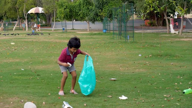 Young volunteer cleaning up garbage to reduce environmental issues - ecology concept. Small Indian kid picking waste material in a garbage bag at a city park - save environment concept. Plogging in...