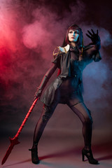 Obraz premium Beautiful leggy busty cosplay girl wearing an erotic leather costume posing with a fake spear on a red background in theatrical smoke.