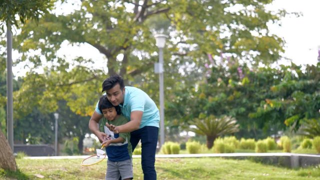 Handsome Indian father teaching his son how to play badminton - outdoor games. A young man teaches his little kid how to hit a shuttle or cock with a racket while playing badminton - fatherhood con...