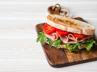 Ham, tomato and arugula sandwich on toasted bread slices on a wood cutting board on wood white background. Copy space