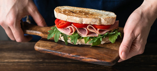Ham, tomato and arugula sandwich on toasted bread slices on a wood cutting board in female hands