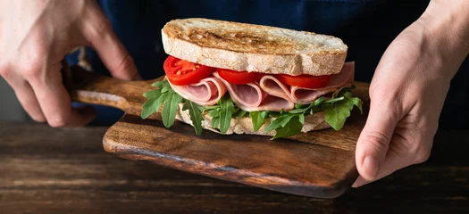 Fototapeten Ham, tomato and arugula sandwich on toasted bread slices on a wood cutting board in female hands © senteliaolga