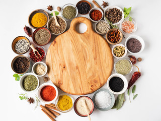 Various dry spices frame flat lay in small bowls and wood cutting board in the center on white wood background. Top view, copy space