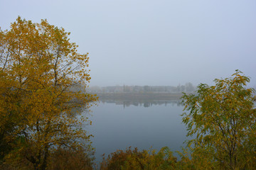 Beautiful landscape, autumnal nature with white dense fog over Career Lake, Dnipro city, Ukraine. Tall trees and large shrubs with yellow leaves around a clean and beautiful lake.