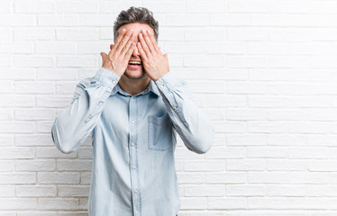 Young handsome man against a bricks wall covers eyes with hands, smiles broadly waiting for a surprise.