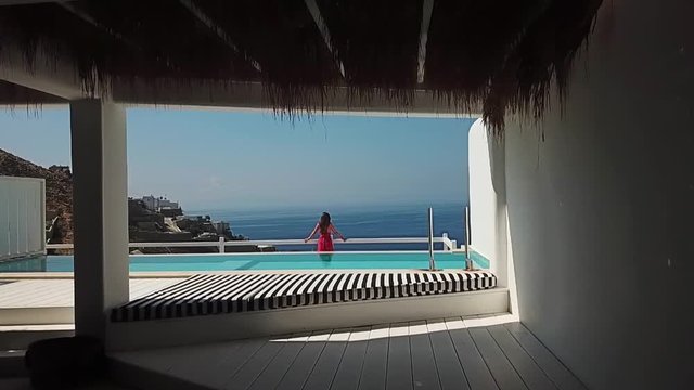 Female in Summer Dress Standing on Terrace by Private Pool in Luxury Resort and Watching Horizon Over Sea, From Interior to Exterior Aerial