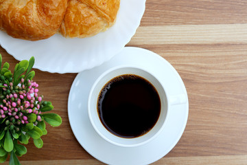 Black coffee in white cup with croissant on wooden background, top view.