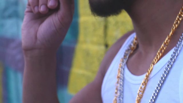 tilt shot following hand going up to gangsters mouth wearing a tank top and chains smoking a spliff in slow motion