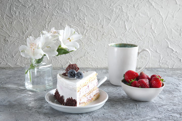 Piece of white mousse dessert cake with blueberries and chocolate on a plate with strawberries, cup of tea and white flowers on grey textured background. Beautiful breakfast. Selective focus