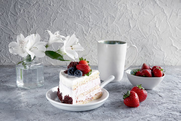 Piece of white mousse dessert cake with blueberries and chocolate on a plate with strawberries, cup of tea and white flowers on grey textured background. Beautiful breakfast. Selective focus