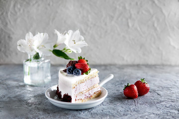 Piece of white mousse dessert cake with blueberries and chocolate on a plate with strawberries and white flowers on light grey textured background. Beautiful breakfast.