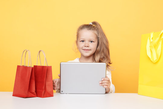Little smiling girl online using laptop and sitting at the desk with shopping bags