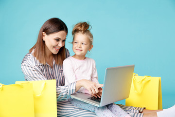 Portrait of mother and daughter with laptop sitting on the floor isolated over the blue studio. Online family shopping concept