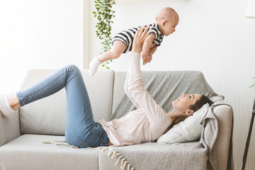 Mother lifting newborn baby in air while lying on sofa