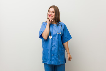 Young nurse woman against a white wall smiling happy and confident, touching chin with hand.