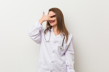 Young doctor woman against a white wall blink at the camera through fingers, embarrassed covering face.