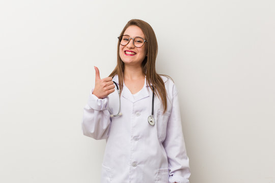 Young doctor woman against a white wall smiling and raising thumb up