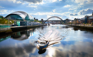 A boat sailing down the River Tyne as it approaches the Millennium Bridge with the Gateshead Sage, Newcastle Crown Court and Newcastle Tyne Bridge in the distance. - obrazy, fototapety, plakaty
