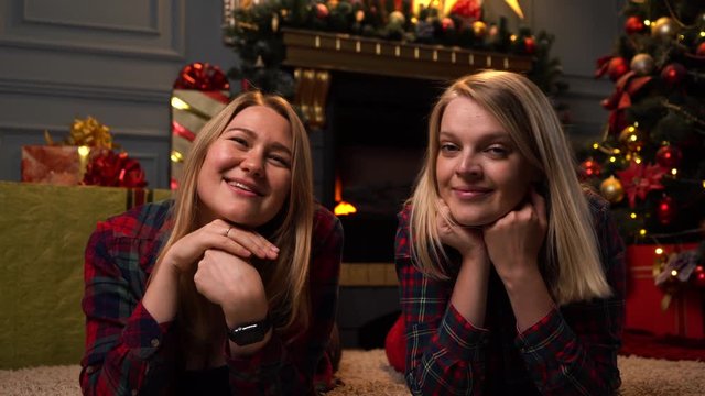 two pretty blondes in red plaid shirts lie on a carpet near a New Year or Christmas tree and look at each other, then into the camera