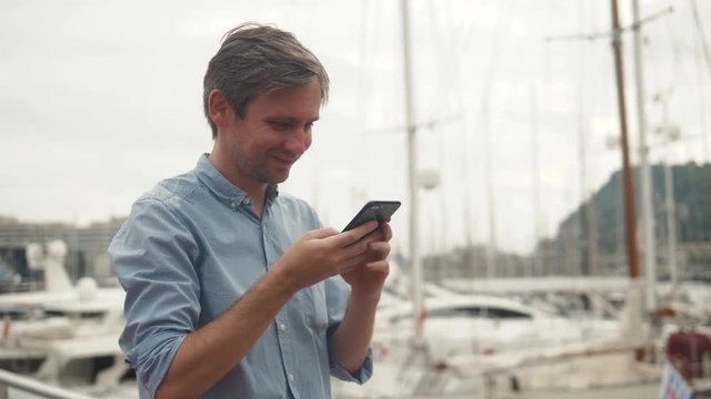 Handsome Caucasian man smiles while using cell phone as he leans against railing at end of breakwater of bay near boats and yachts parking