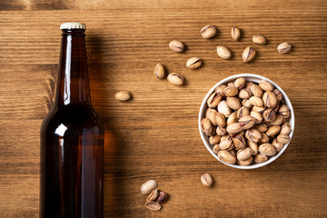 A bottle of dark beer and fried salted pistachios in a white bowl on a wooden background, top view, flat lay.