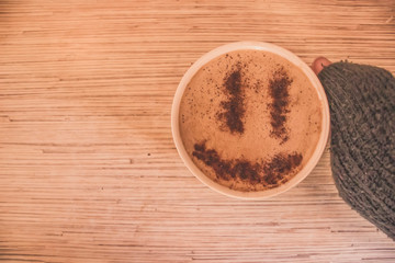Mug of cappuccino with cinnamon for breakfast.Cinnamon powder in form of a smiley face on coffee foam.Morning cup coffee with hot cappuccino in hand.Hand in a sweater holding mug of coffee with foam.