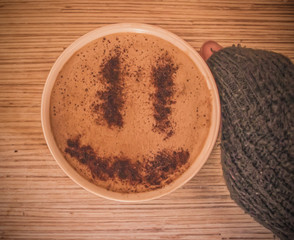 Mug of cappuccino with cinnamon for breakfast.Cinnamon powder in form of a smiley face on coffee foam.Morning cup coffee with hot cappuccino in hand.Hand in a sweater holding mug of coffee with foam.