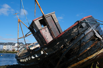 Old wooden shipwreck at the edge of the Corrib River as it flows into Galway Bay, Ireland showing the decayed wood, and houses in the background, taken on a sunny summer day.