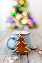Merry Christmas! Christmas tree-shaped gingerbread cookie near cups of cocoa and marshmallows. Beautiful blurred christmas tree background with luminous garland.